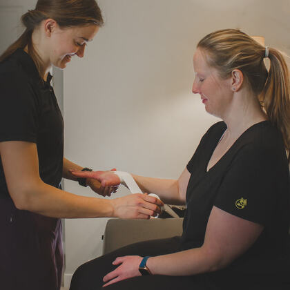 Erin is standing in front of an athlete who is sitting on her treatment table. She is applying a prophylactic taping technique to support the athlete's wrist.