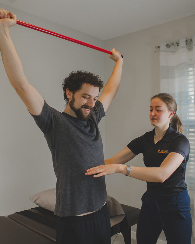 Erin is standing next to a client, walking them through an overhead squat movement. Her aim is to help correct which muscles are working, and help cue the client on how to achieve a coordinated movement. Ultimately this will help enhance postural control, core strength, muscle flexibility and joint health.