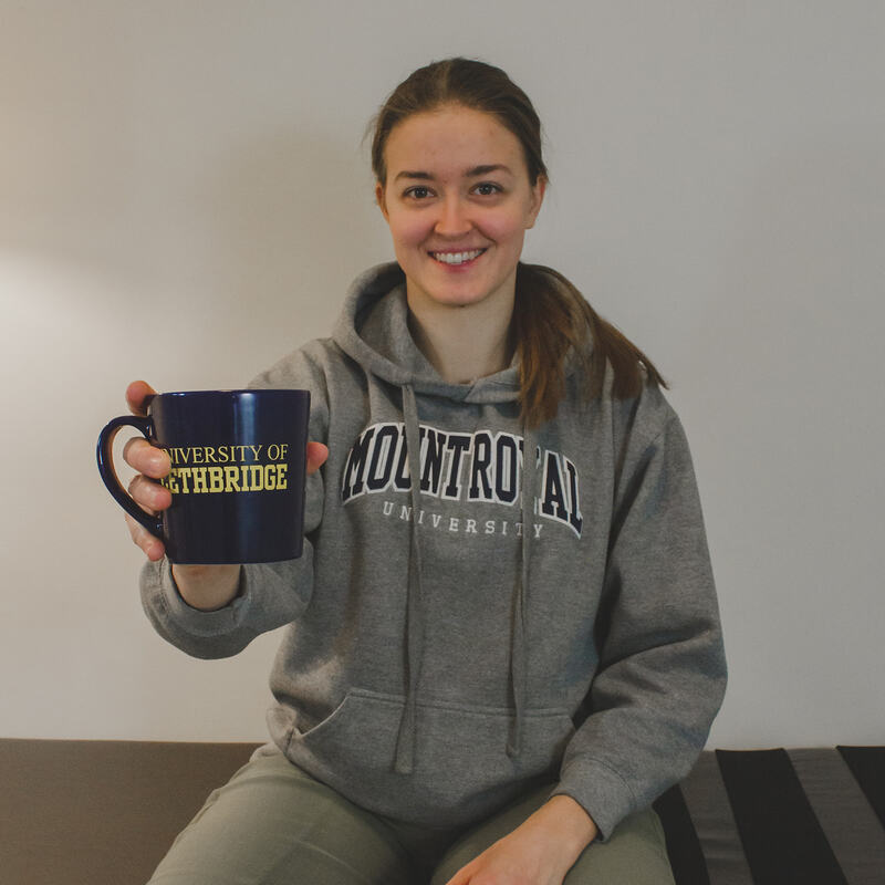 Erin is smiling while wearing a hoodie from Mount Royal University and holding a mug from the University of Lethbridge.